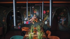 Um restaurante com uma longa mesa de madeira, cadeiras estofadas verdes e um mural com anjos e frutas.