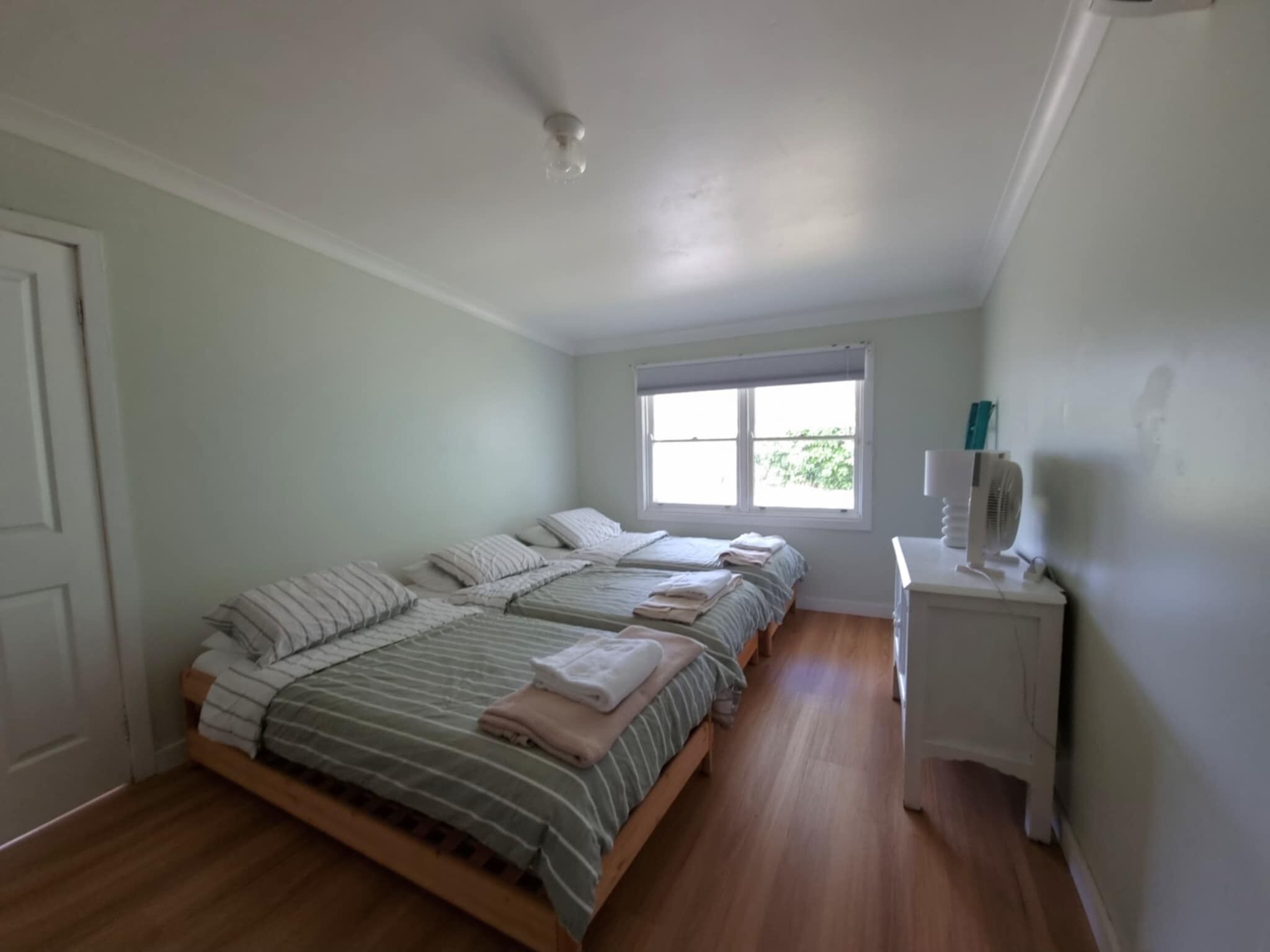 Une chambre avec deux lits, une fenêtre, une table de chevet avec un ventilateur et une porte.