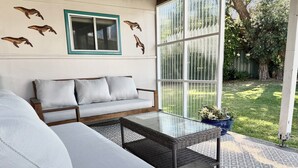 Uma área de estar moderna ao ar livre, com um sofá e uma mesa com tampo de vidro, uma janela com vista para o jardim e arte decorativa na parede.