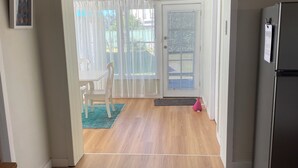 Quarto espaçoso com piso de madeira, geladeira, mesa de jantar com cadeiras e vista para área externa através de uma porta de vidro.