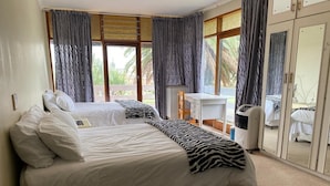 Une chambre avec un grand lit, un bureau et une chaise. Les fenêtres sont dotées de rideaux, et une porte-fenêtre coulissante donne sur un balcon.