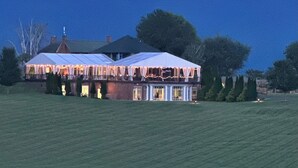 A large, well-lit building with multiple covered areas, surrounded by a grassy field and trees, with a hill in the background. - Libby's Lake View on Smith Mountain Lake  - Inquire about long term rentals! (Moneta)