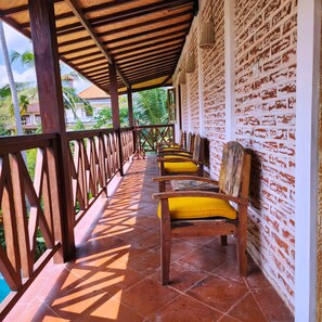 Un espacio de descanso techado al aire libre con sillas de madera y cojines amarillos, suelo de baldosas y pared de ladrillo con un diseño blanco.