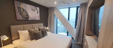 A modern hotel room with a large bed, a ceiling fan, a painting on the wall, and a view of the city through the windows.