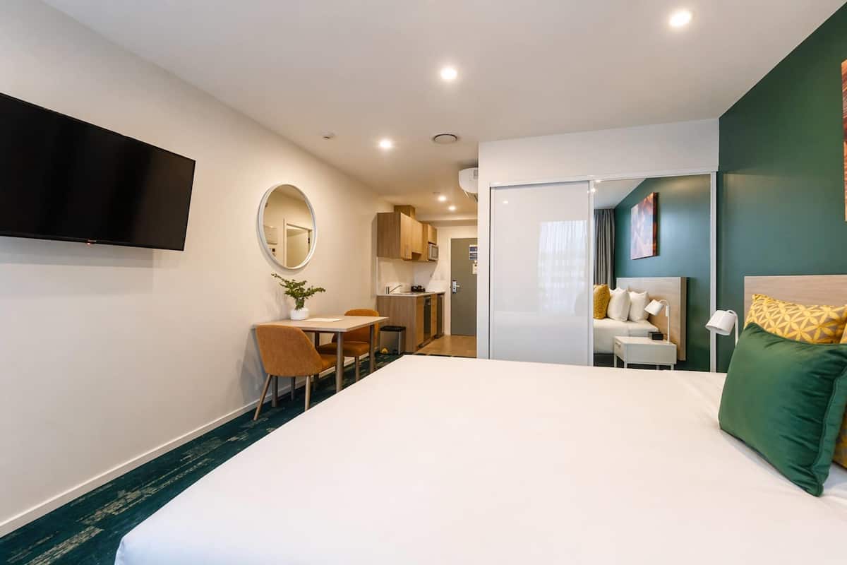 Ein Hotelzimmer mit Bett, Schreibtisch mit Stuhl, Wandfernseher, einer kleinen Küchenzeile und einem durchgegangenen Badezimmer.
