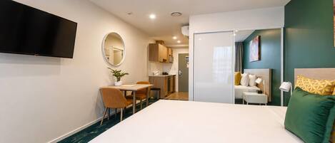 Habitación de hotel con cama, escritorio con silla, televisión en la pared, pequeña cocina y baño visible a través de una puerta abierta.