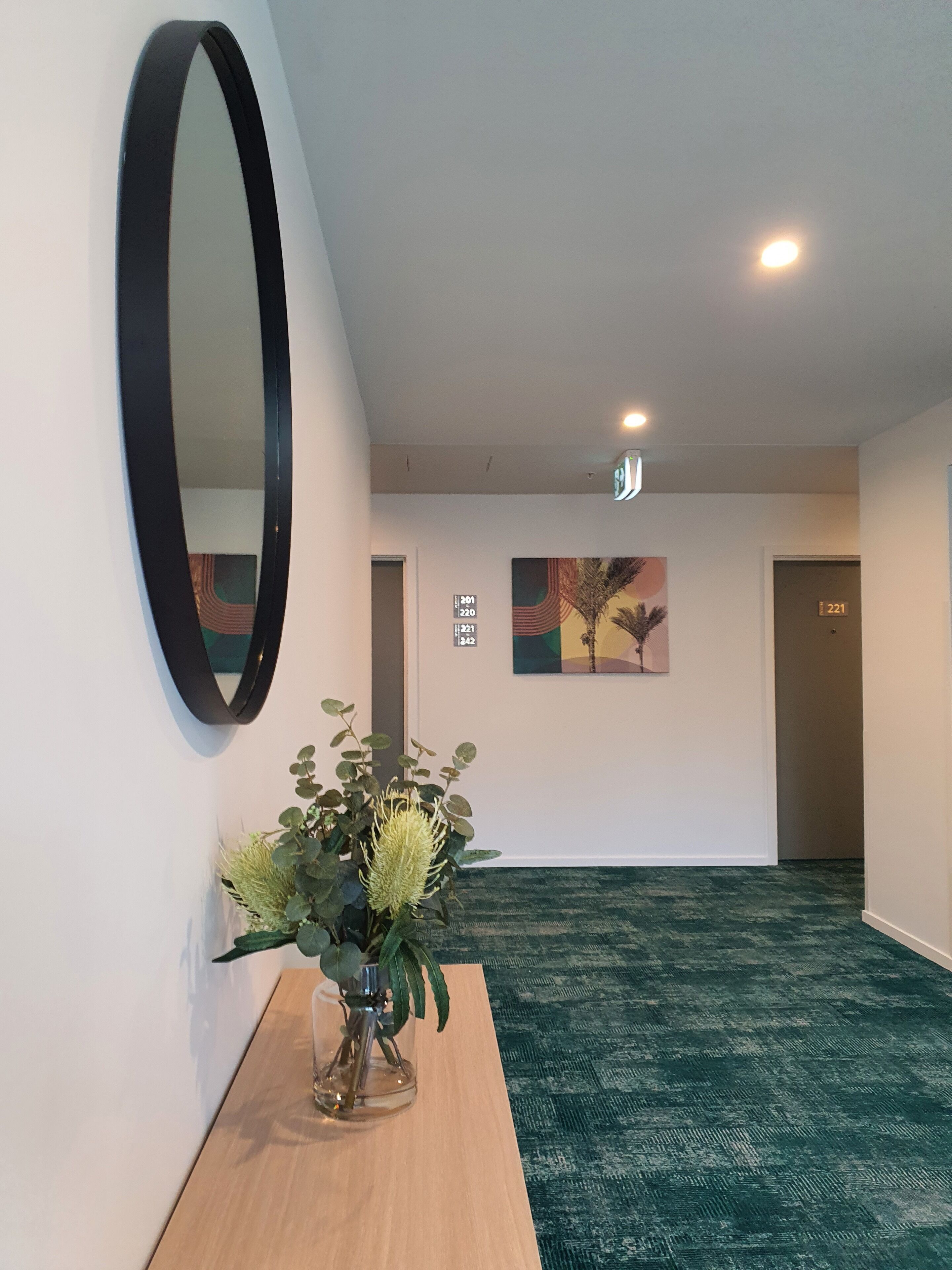 A hallway with a mirror, a potted plant, a painting, and numbered doors.