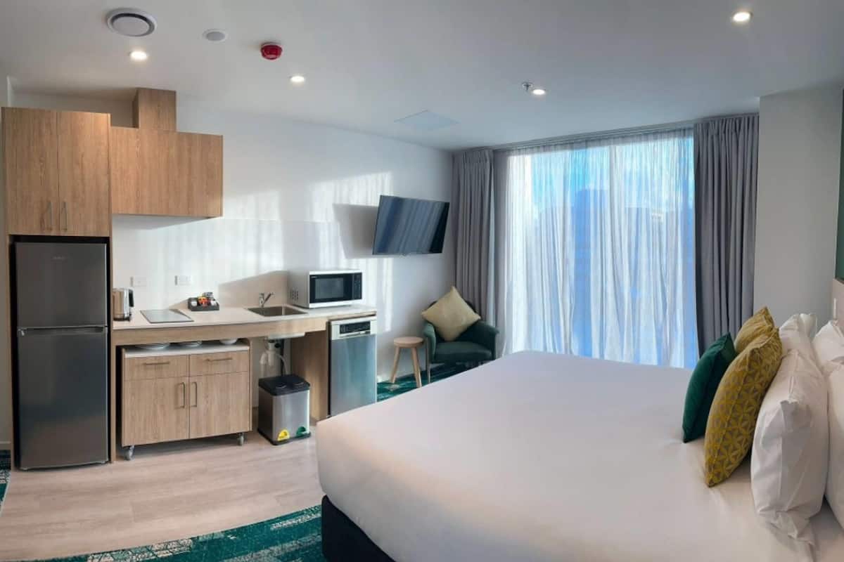 Ein Hotelzimmer mit einem Bett, einem Schreibtisch mit Computer, einem Fernseher und einer Küchenzeile mit Kühlschrank sowie einem Fenster mit Vorhängen.