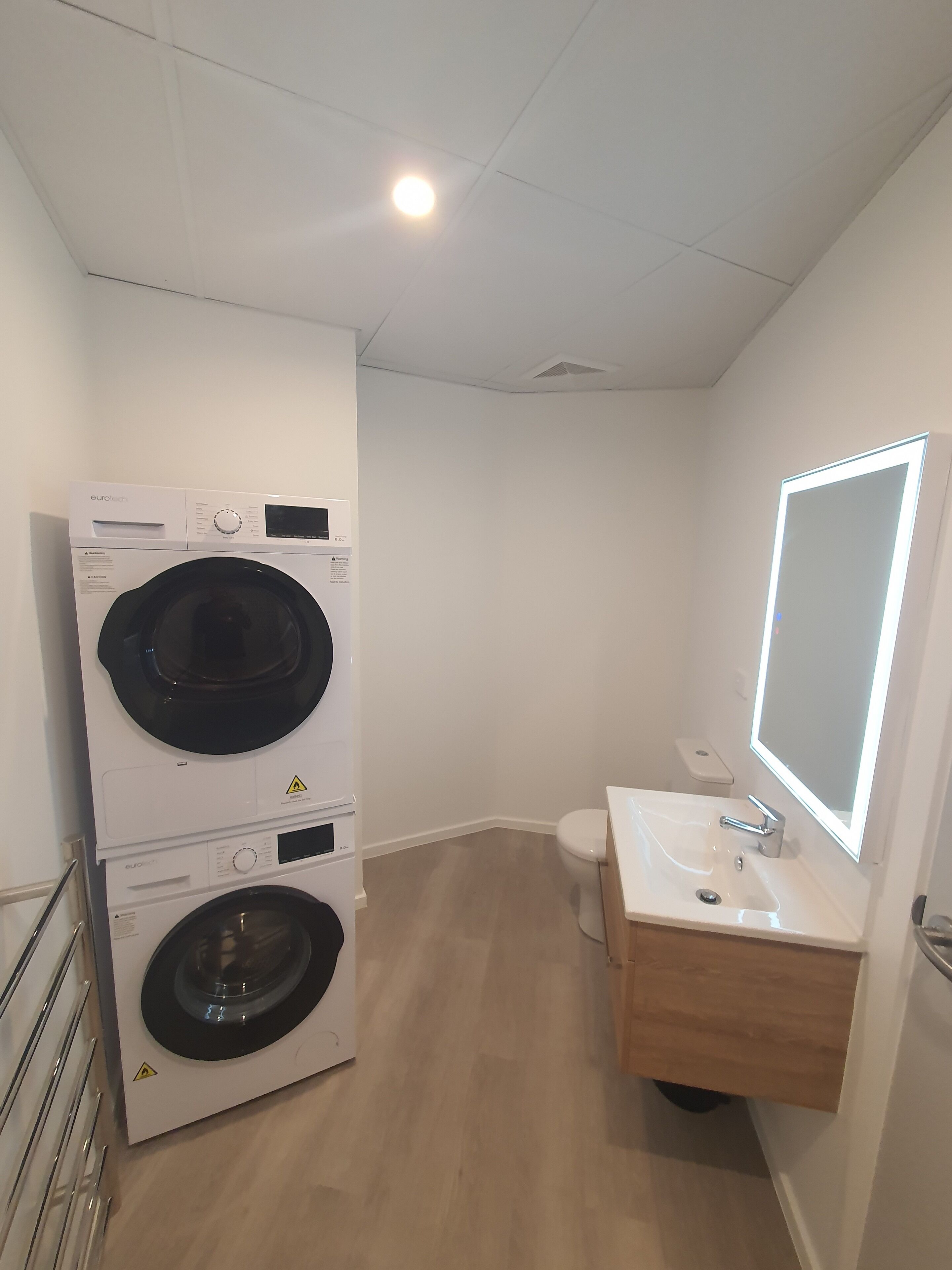 A compact bathroom with a washing machine, toilet, and a mirror with built-in lighting.
