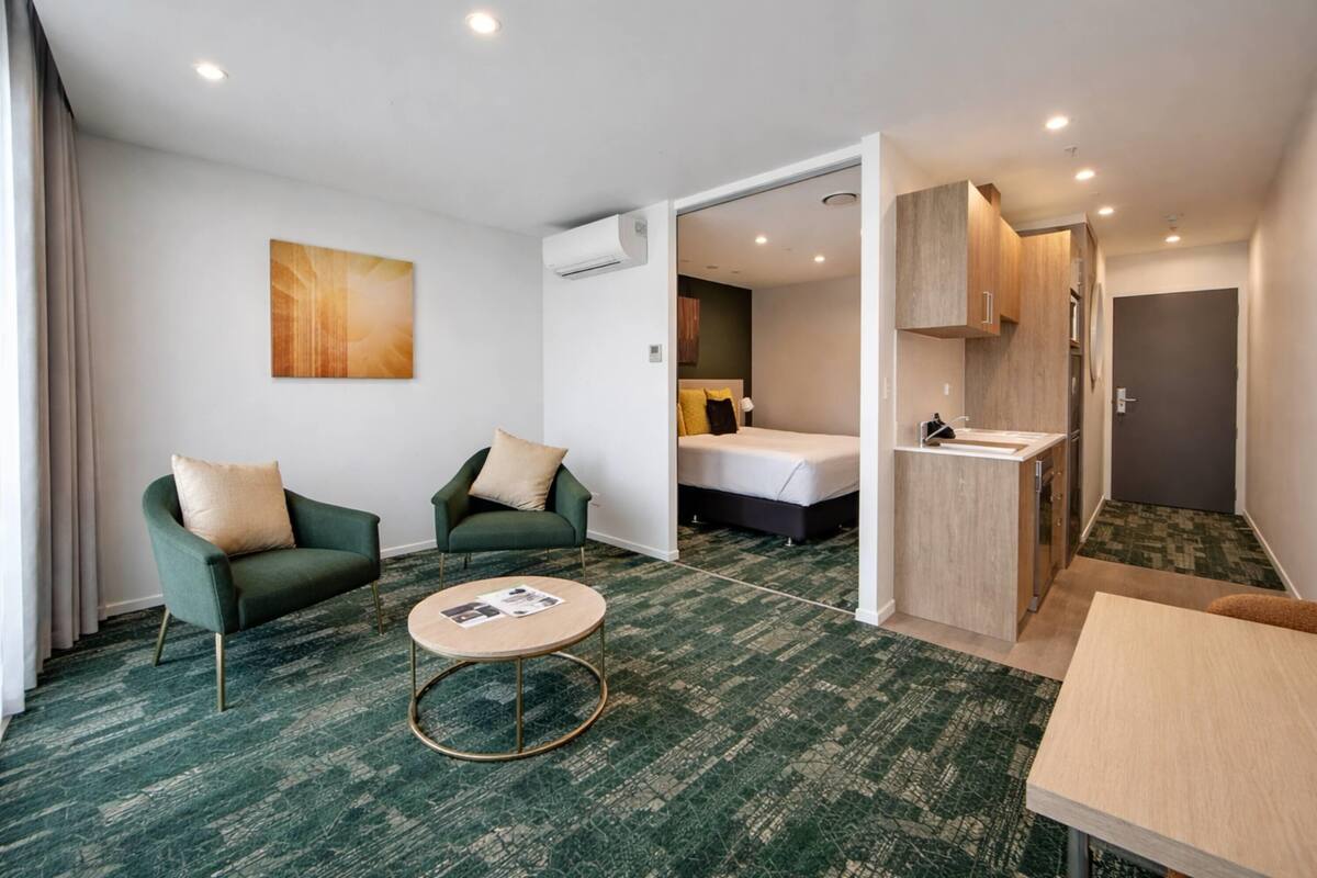 Ein Hotelzimmer mit einem Bett, zwei Sesseln, einem runden Couchtisch, einer Küchenzeile und einem Bild an der Wand.