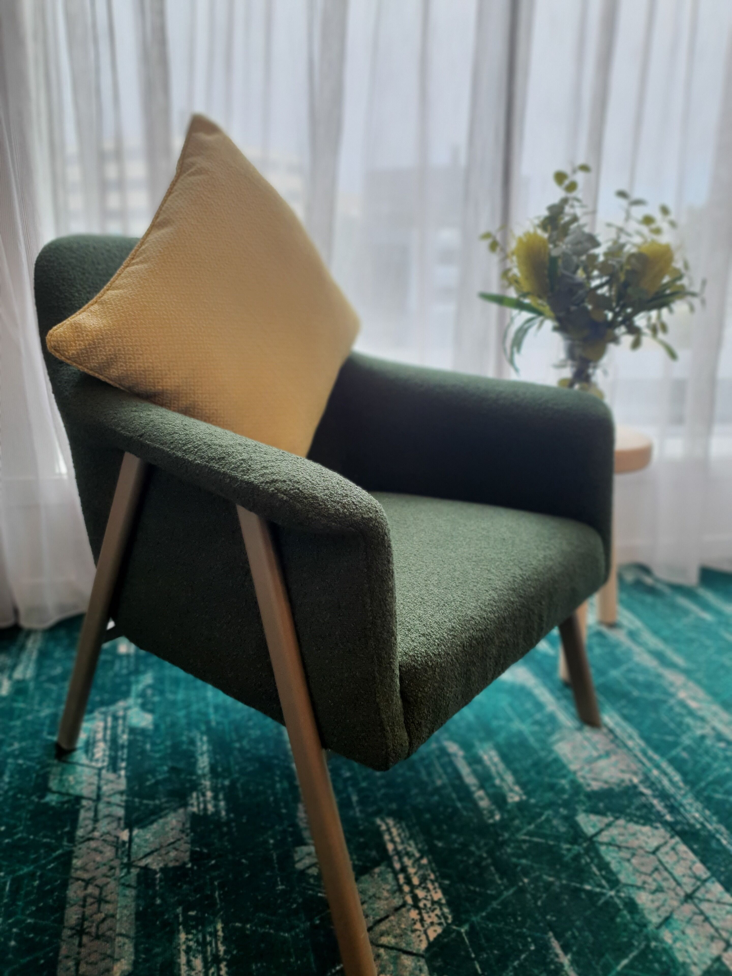 Ein grüner Sessel mit einem beigen Kissen, vor einem Fenster mit transparenten Vorhängen und einer Vase mit gelben Blumen platziert.