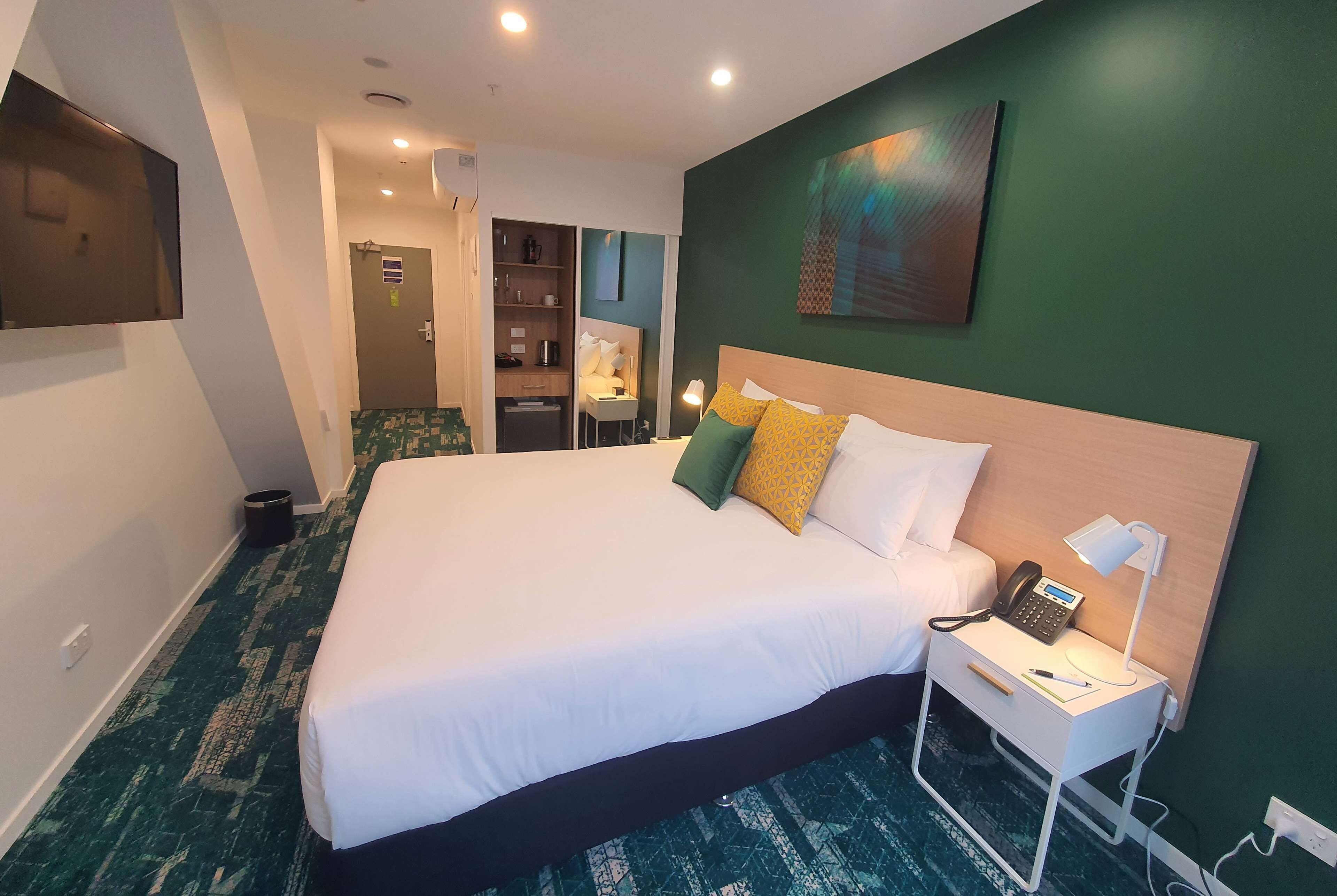 A hotel room with a large bed, a bedside table with a lamp, a phone, and a small desk with a chair. There is a green accent wall and a painting above the bed.