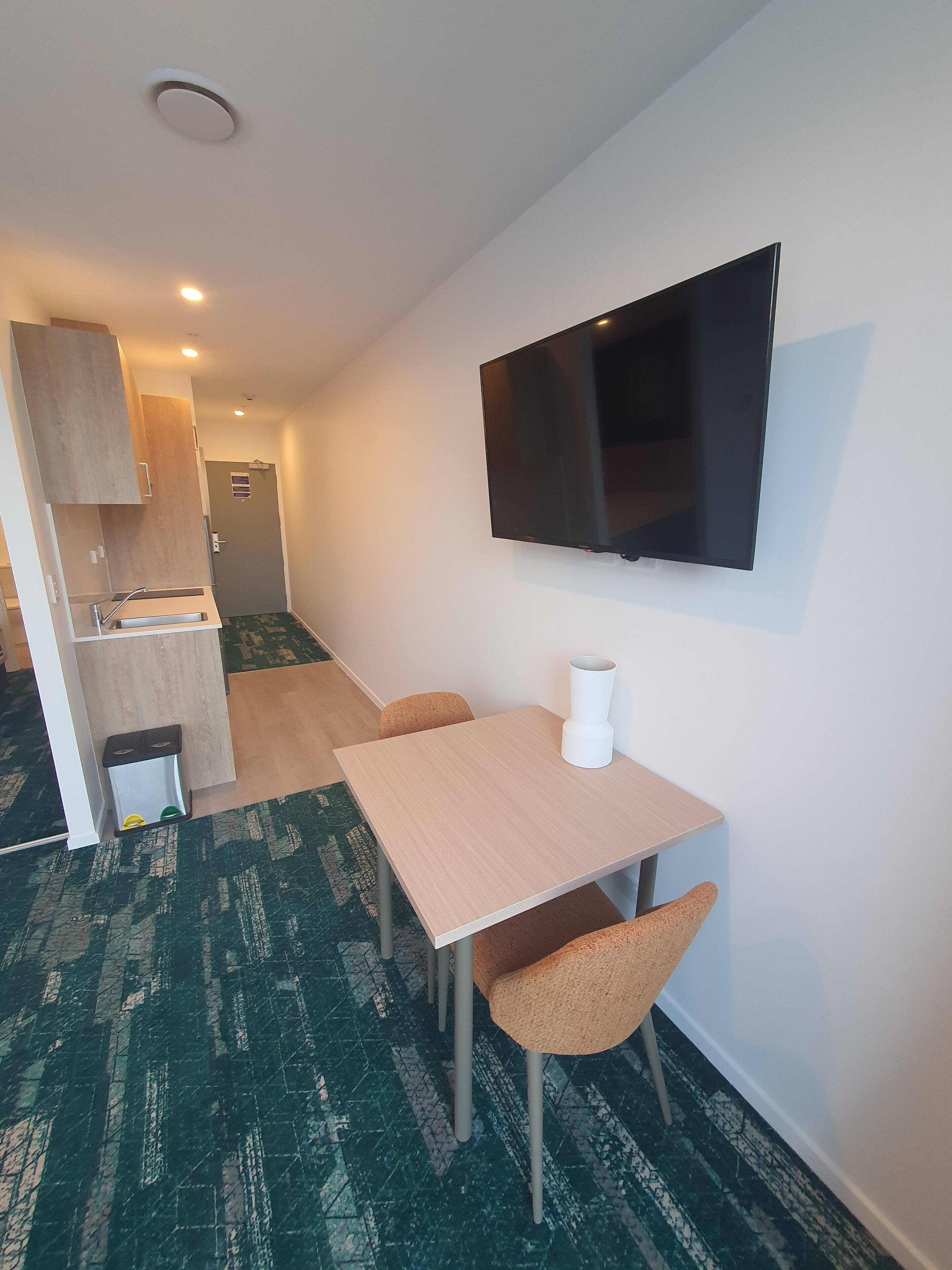 A compact hotel room with a kitchenette, a flat-screen TV mounted on the wall, a small table with two chairs, and a trash bin.