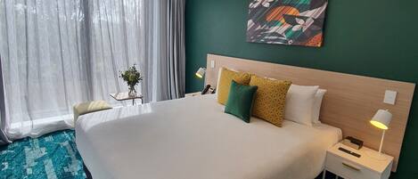 A hotel room with a large bed, a bedside table with a lamp, a green accent wall, and a window with sheer curtains.