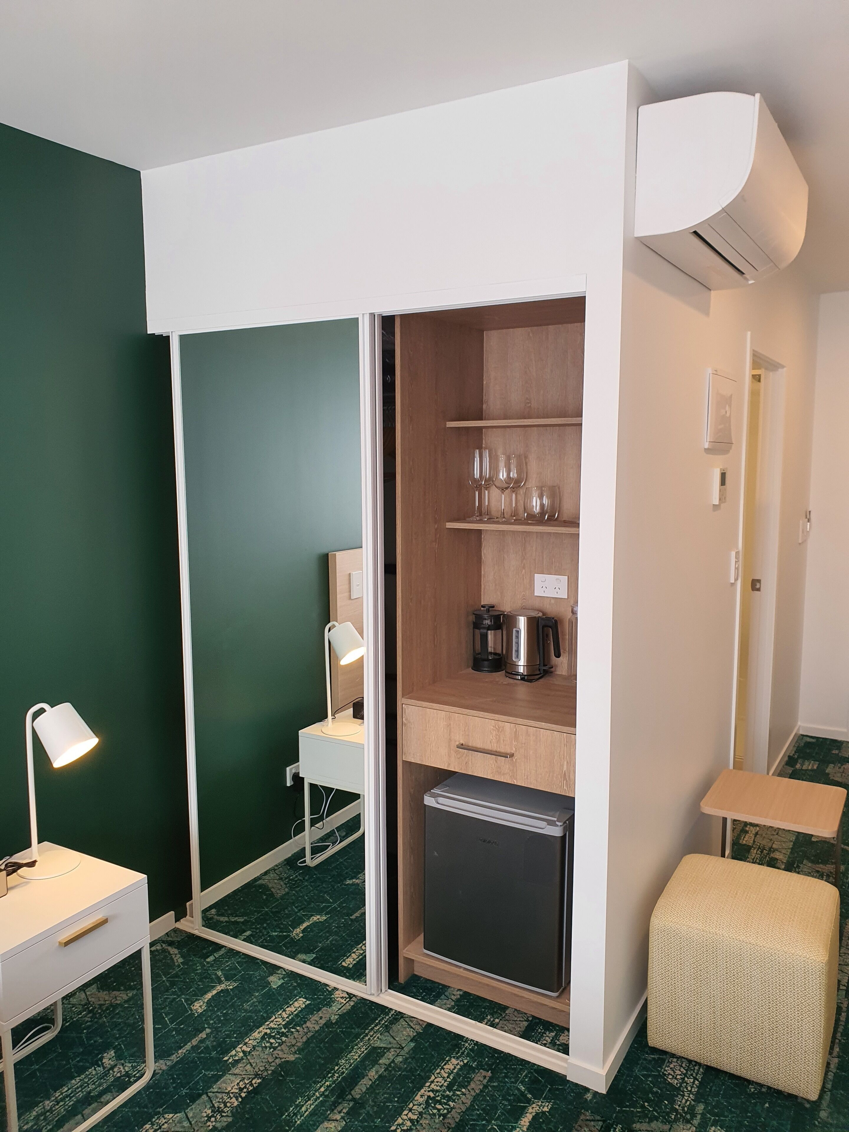 Una habitación compacta con kitchenette, un escritorio con lámpara, un taburete pequeño y un armario con puerta corredera.