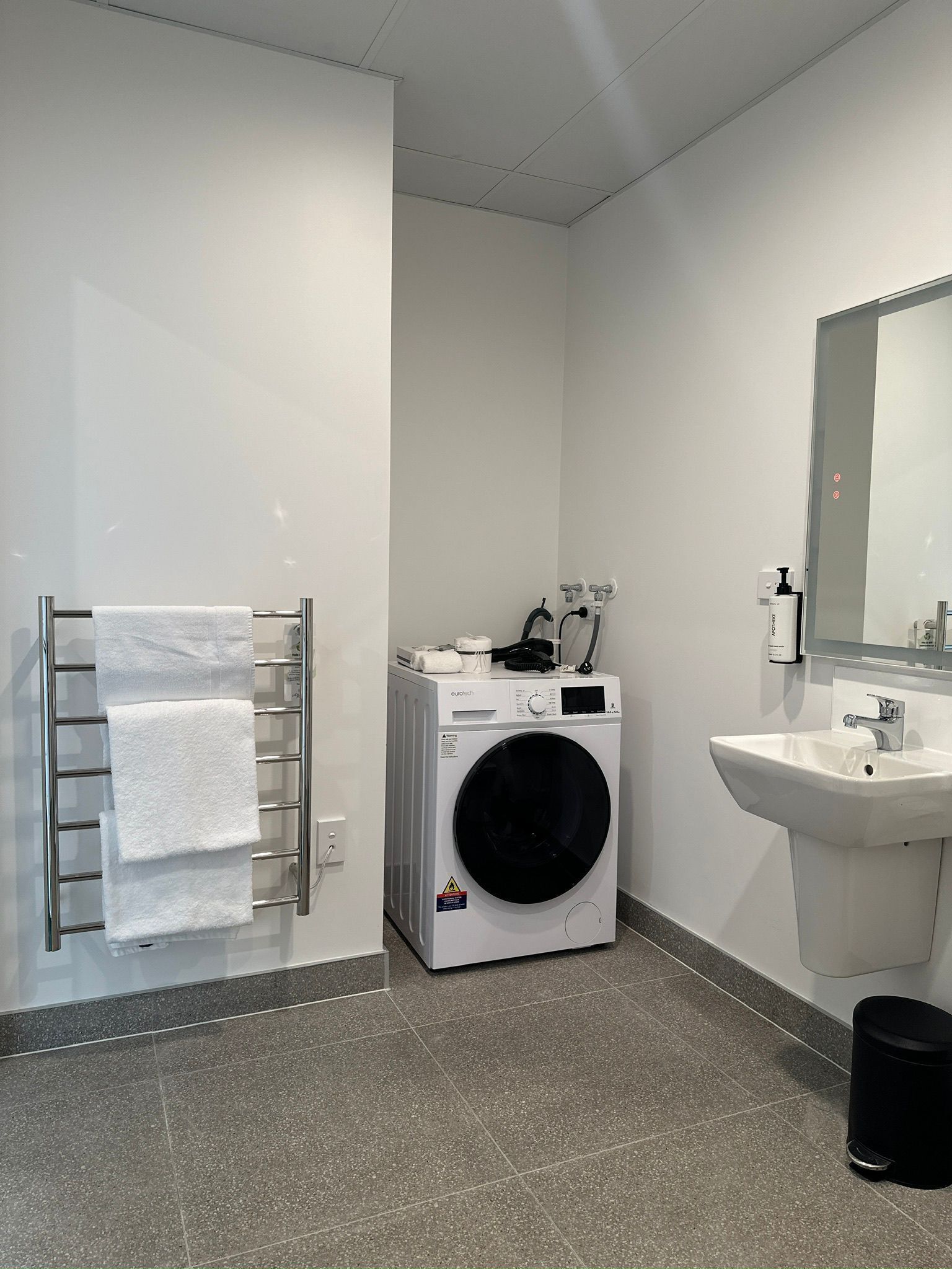 A modern bathroom with a built-in washing machine, a towel rack, a sink, and a trash bin.
