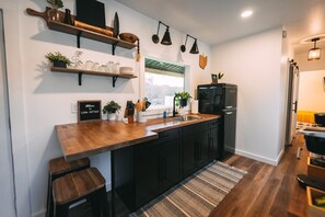 Una cocina moderna con encimeras de madera, armarios negros y un refrigerador negro. Hay repisas con cristalería y plantas, y una ventana que proporciona luz natural.