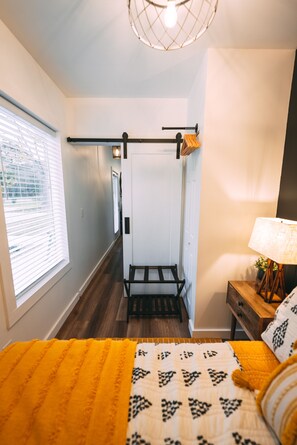 Un dormitorio con cama, mesita de noche, lámpara, ventana con persianas y un pasillo que lleva a otra habitación.