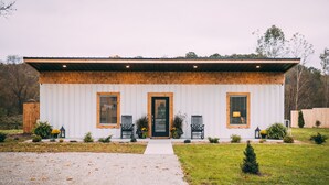 Une maison de plain-pied à l’extérieur blanc et à la façade en bois, dotée d’une porte d’entrée, de deux fenêtres et d’un petit porche avec deux chaises et des plantes en pot.