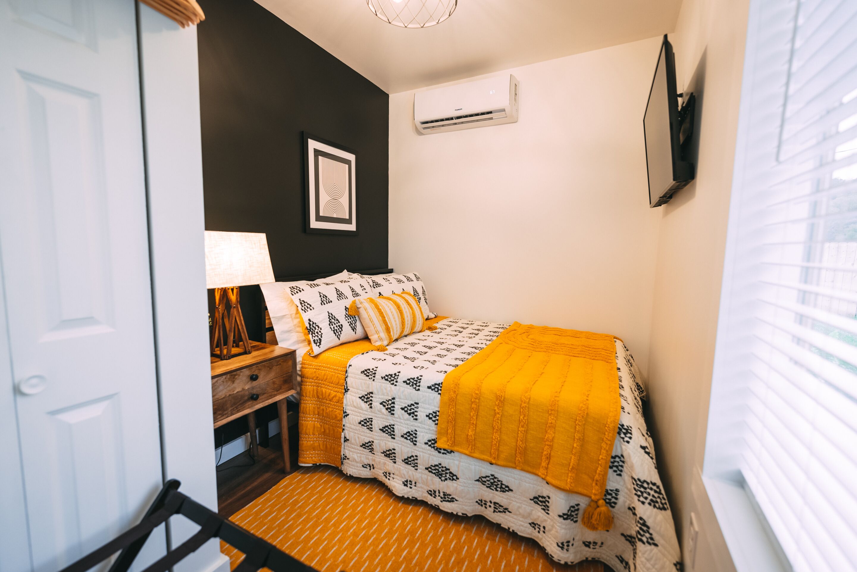 Un dormitorio con cama, una mesita de noche, una lámpara, aire acondicionado instalado en la pared y un cuadro enmarcado.