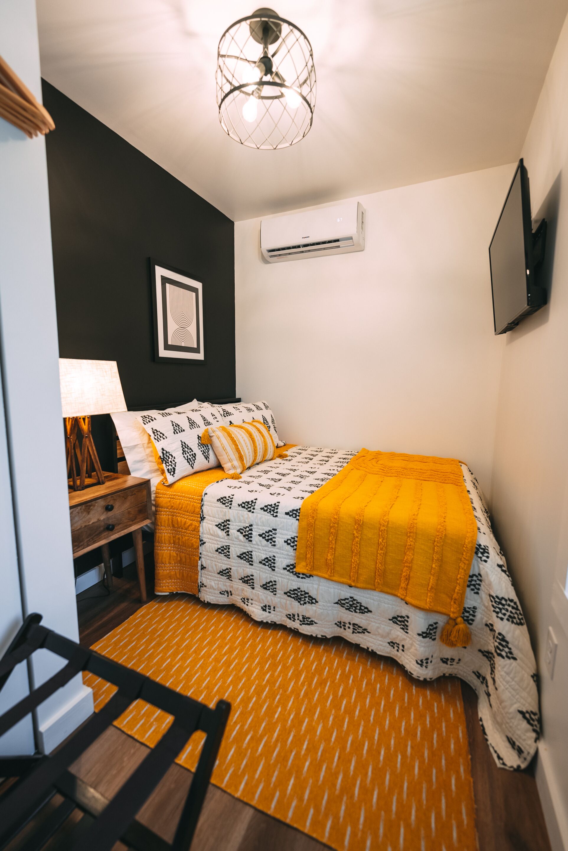 A bedroom with a bed, a bedside table, a lamp, an air conditioner, and a framed picture on the wall.