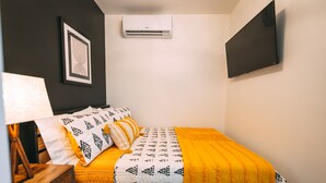 Une chambre à coucher comprenant un lit, une table de chevet, une lampe, un appareil de climatisation et un tableau encadré accroché au mur.