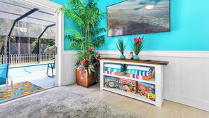 Een kamer met een zwembad, een grote plant, een televisie en een plank met gezelschapsspellen.