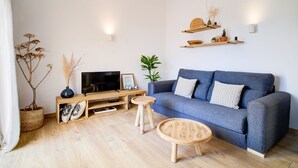 A modern living room with a grey sofa, wooden coffee table, and a TV on a wooden stand. There are decorative shelves above the sofa and a potted plant on the floor. - Surmar - Apartment with pool in Cap d'Artrutx (Ciutadella de Menorca)