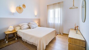 A bedroom with a bed, bedside lamps, a wooden dresser, and a window with sheer curtains. - Surmar - Apartment with pool in Cap d'Artrutx (Ciutadella de Menorca)