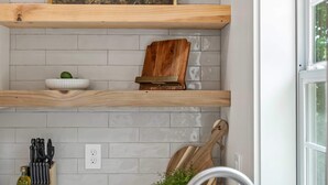Une cuisine avec un plan de travail blanc, un évier en acier inoxydable, une planche à découper en bois, un range-couteaux, une bouteille d’huile d’olive, une plante en pot et un tableau encadré accroché au mur.
