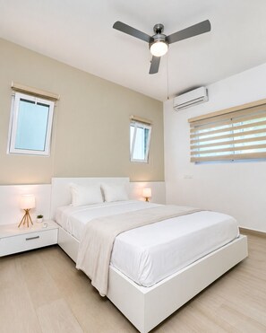 A bedroom with a bed, bedside tables, a ceiling fan, and windows. - 2JA Management Group - White Hill Condos (Sosúa)