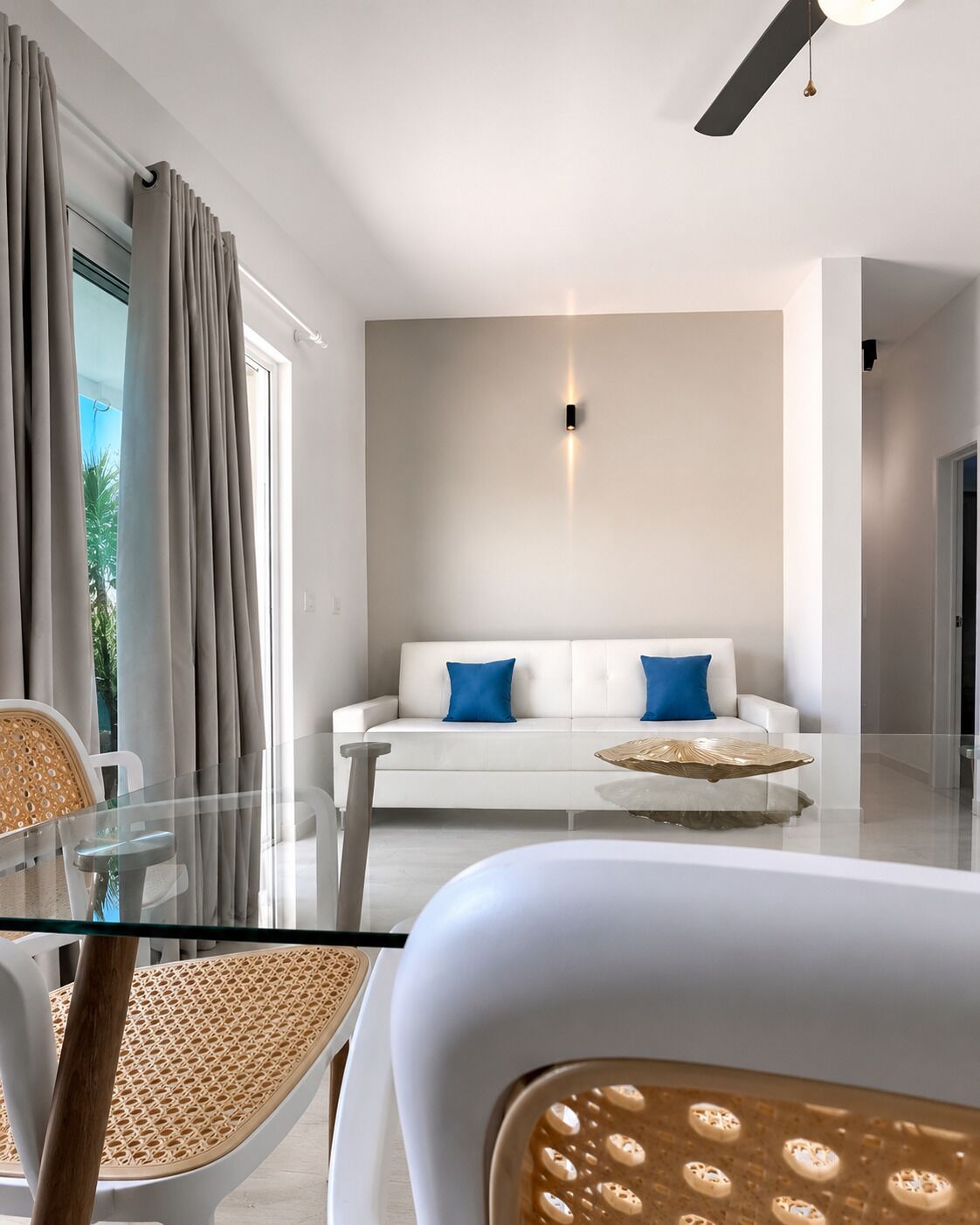 Una sala de estar moderna con una mesa de centro de vidrio, sofá blanco con cojines azules y un ventilador de techo.