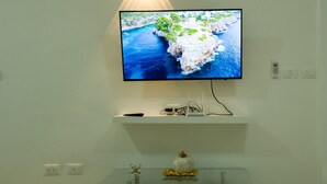 Un televisor de pantalla plana montado en una pared blanca, con una lámpara en el techo arriba. Debajo del televisor, hay una mesa de centro de vidrio con objetos decorativos.