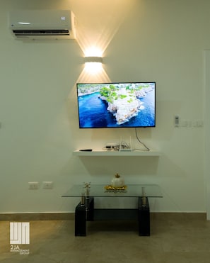A flat-screen TV mounted on a white wall, with a ceiling light above it. Below the TV, there is a glass coffee table with decorative items.