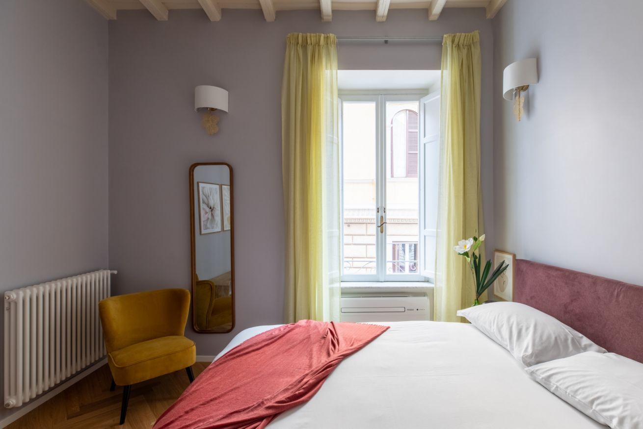 A bedroom with a bed, a chair, a radiator, a window with curtains, and a wall-mounted lamp.