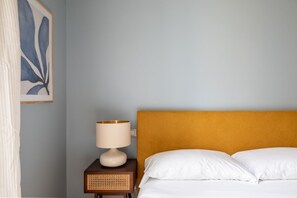 A bedroom with a bed, white pillows, a wooden nightstand with a lamp, and a framed artwork on the wall.