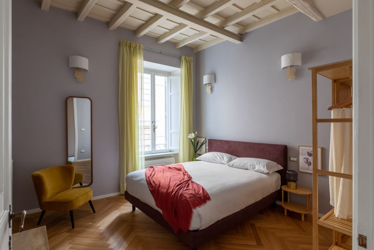 A bedroom with a bed, a chair, a window with curtains, and a mirror on the wall.