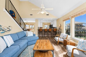 A spacious living area with a blue sofa, wooden coffee table, wicker chairs, and a view of a water body through large windows.