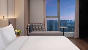 Une chambre d’hôtel moderne, dotée d’un grand lit, d’un canapé, d’une petite table et offrant une vue sur le paysage urbain grâce à une grande fenêtre.