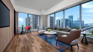 Un salon moderne comprenant un canapé, des fauteuils, une table basse et un téléviseur. De grandes fenêtres offrent une vue sur la ville.
