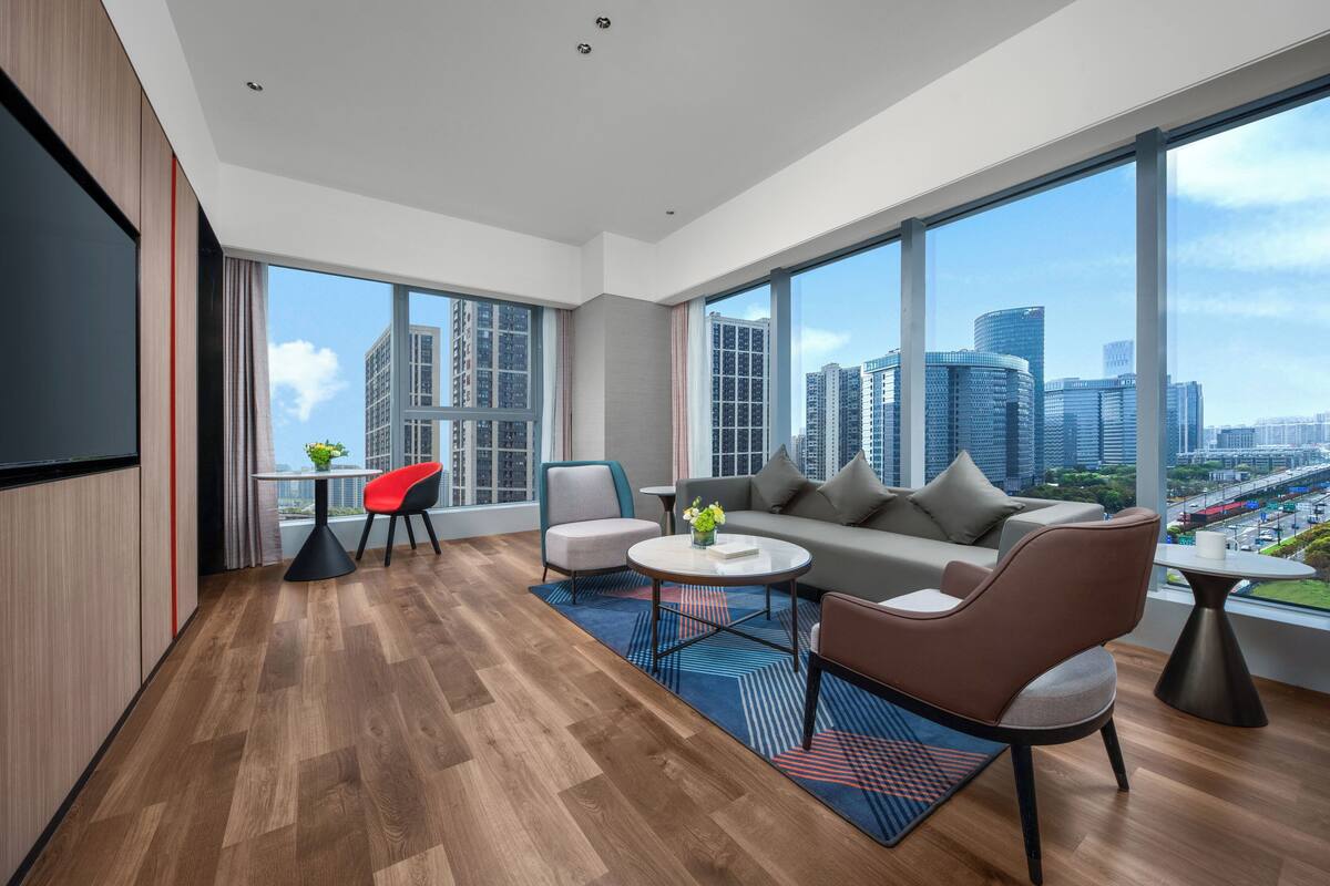Un salon moderne comprenant un canapé, des fauteuils, une table basse et un téléviseur. De grandes fenêtres offrent une vue sur la ville.