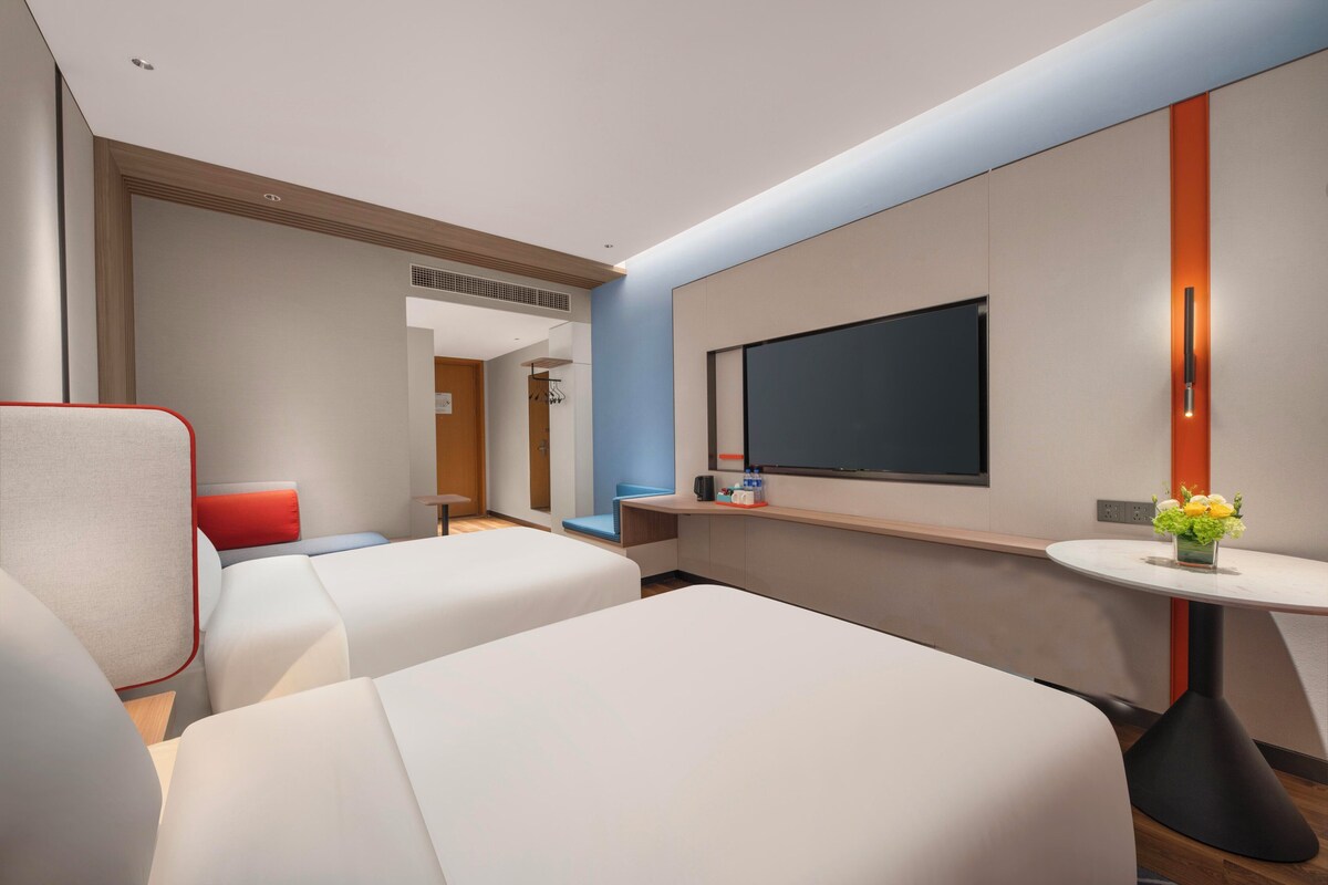 Une chambre d’hôtel moderne avec deux lits, une télévision, une petite table avec un vase et une chaise rouge.