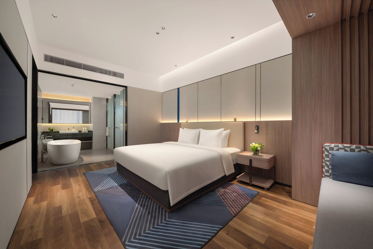 Une chambre d’hôtel moderne, dotée d’un grand lit, d’une table de chevet avec une lampe, d’un petit coin salon et d’une salle de bain visible à travers une porte ouverte.
