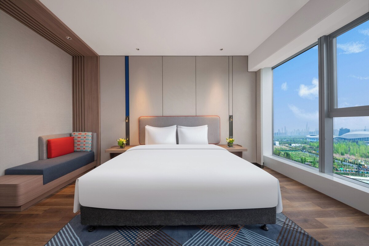 Une chambre d’hôtel moderne avec un grand lit, un coin salon agrémenté d’un coussin rouge, une table de chevet avec un vase et une grande fenêtre donnant sur la ville.