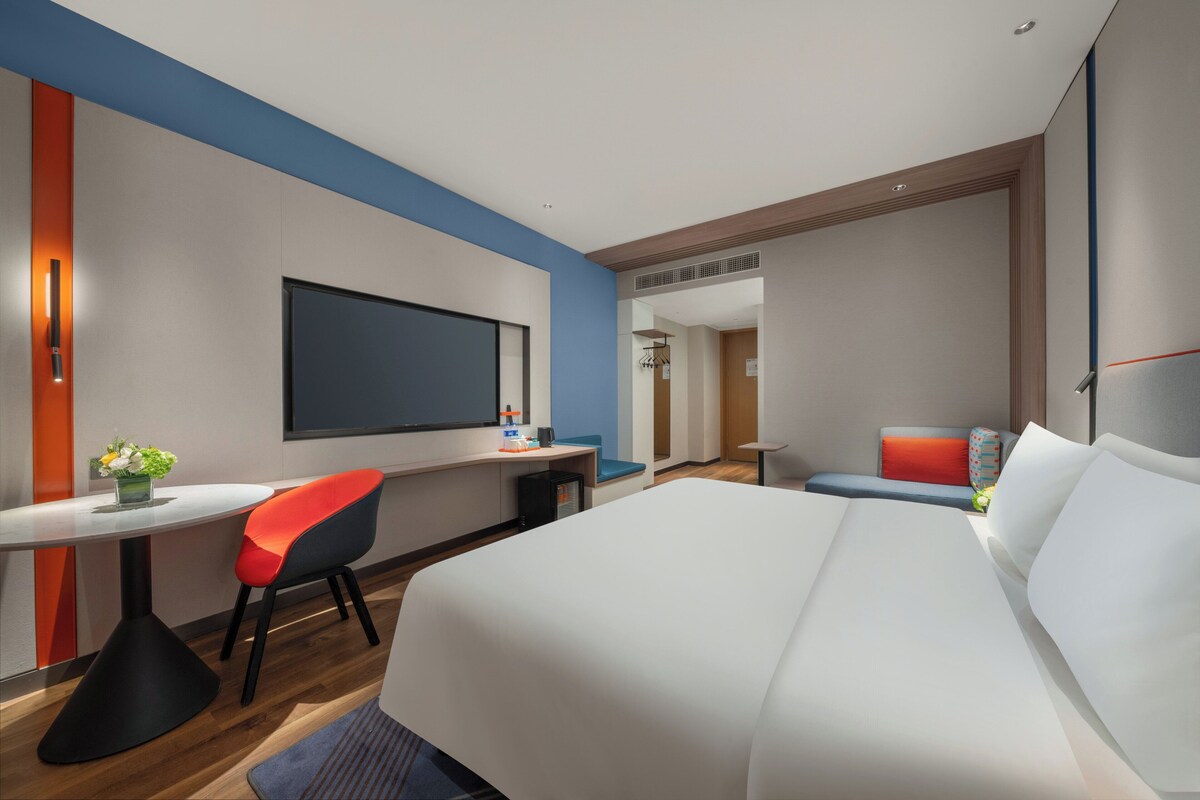 Une chambre d’hôtel moderne dotée d’un grand lit, d’un bureau avec une chaise, d’une télévision et d’un coin salon avec un canapé et des fauteuils.