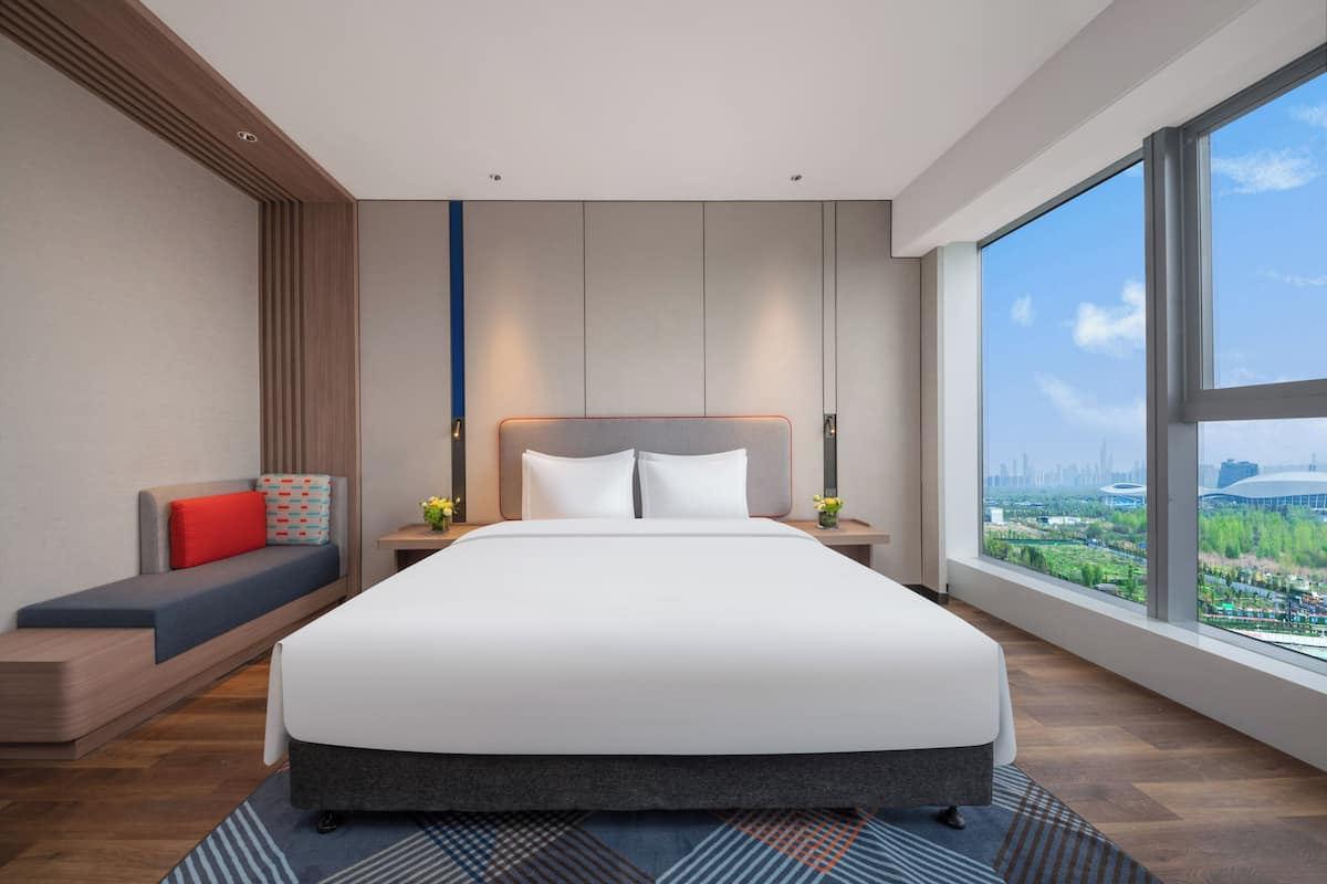 Une chambre d’hôtel moderne avec un grand lit, un coin salon agrémenté d’un coussin rouge, une table de chevet avec un vase et une grande fenêtre donnant sur la ville.