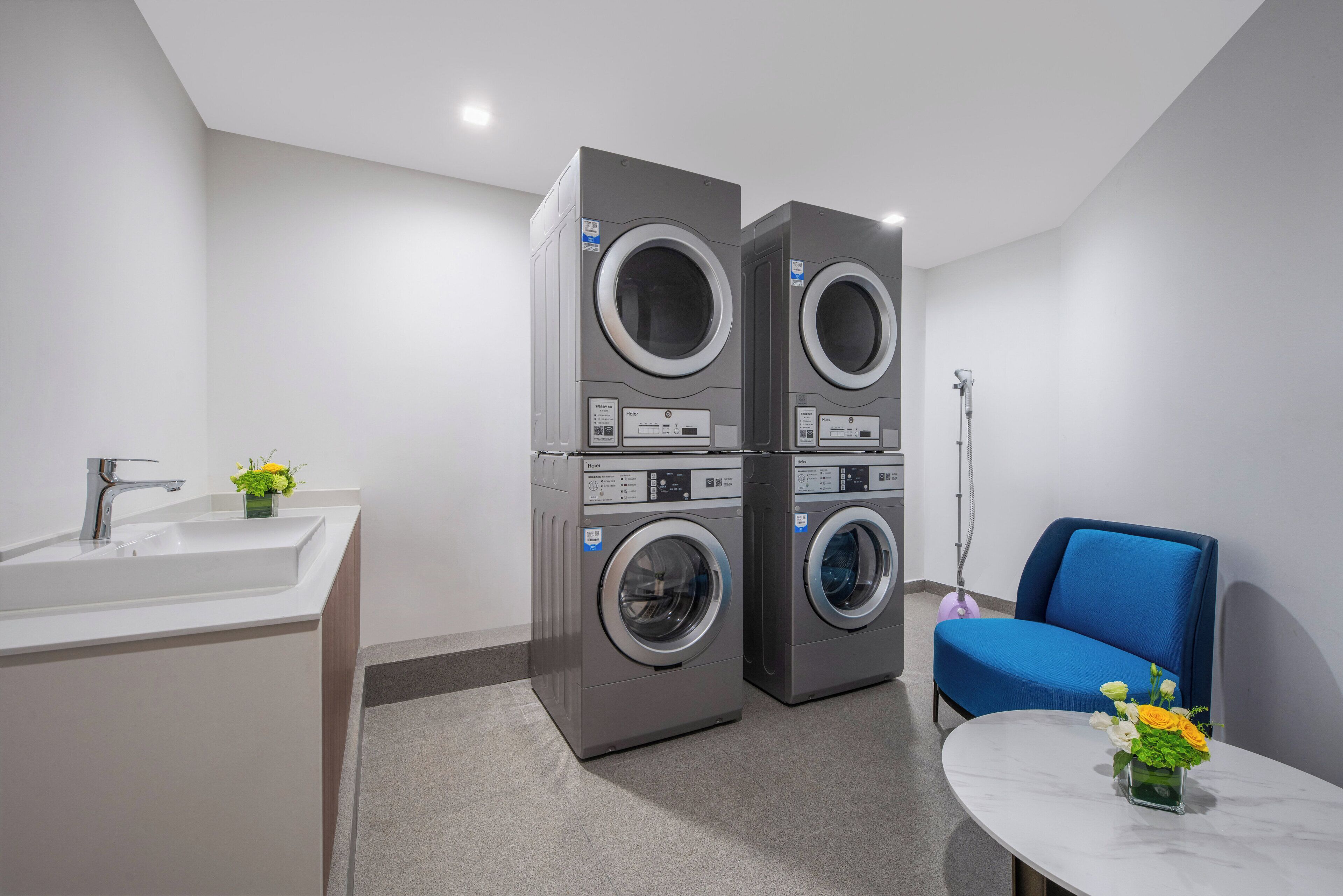 A modern laundry room with two stacked front-loading washing machines, a sink, and a blue chair with a vase of flowers.