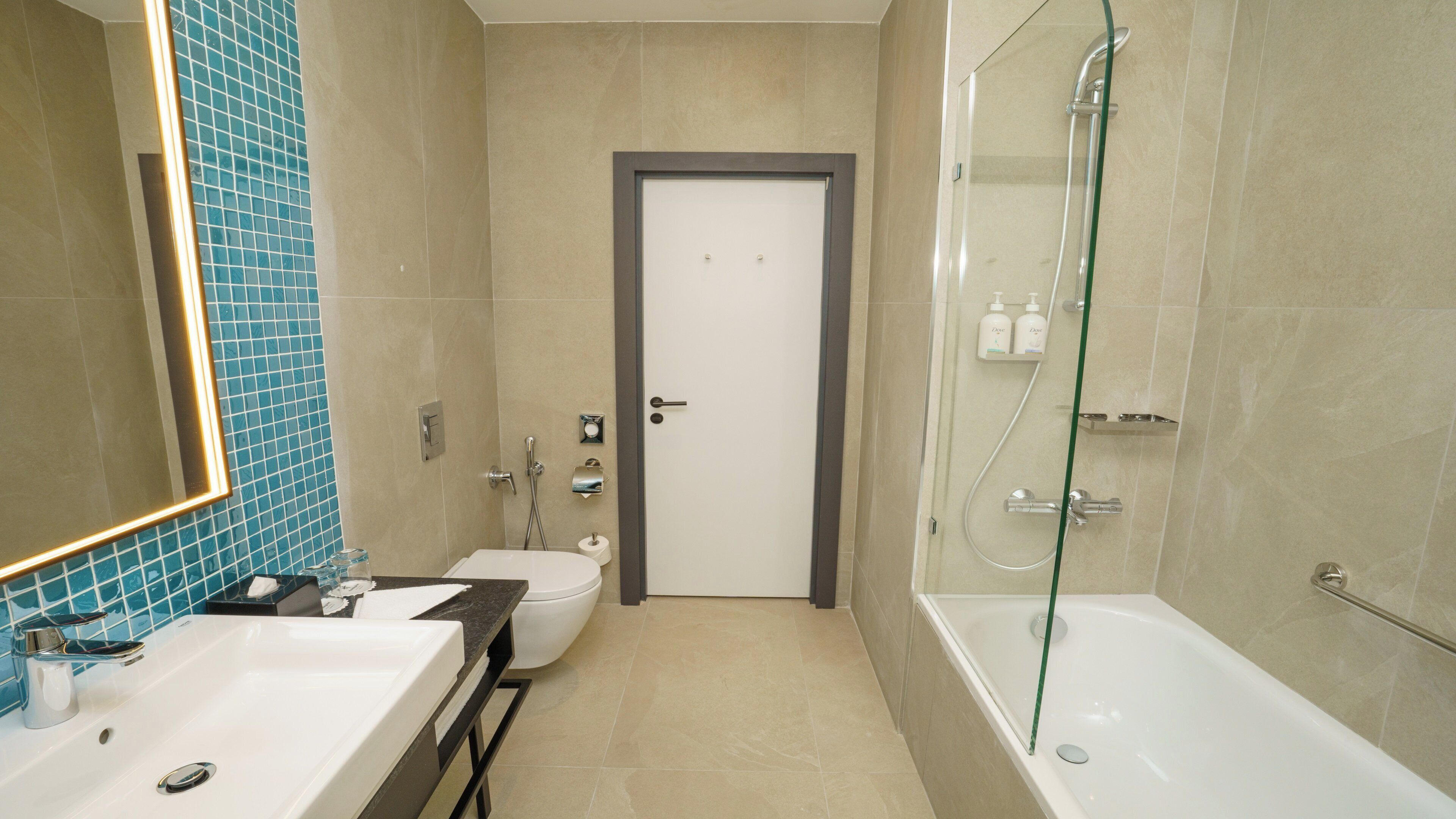 Une salle de bain avec une baignoire blanche, un lavabo et des toilettes. On y trouve une porte surmontée d’un miroir, ainsi qu’un espace douche doté d’une porte en verre.