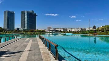 Um calçadão à beira-mar com uma piscina azul límpida, um deck de madeira e edifícios altos modernos ao fundo.