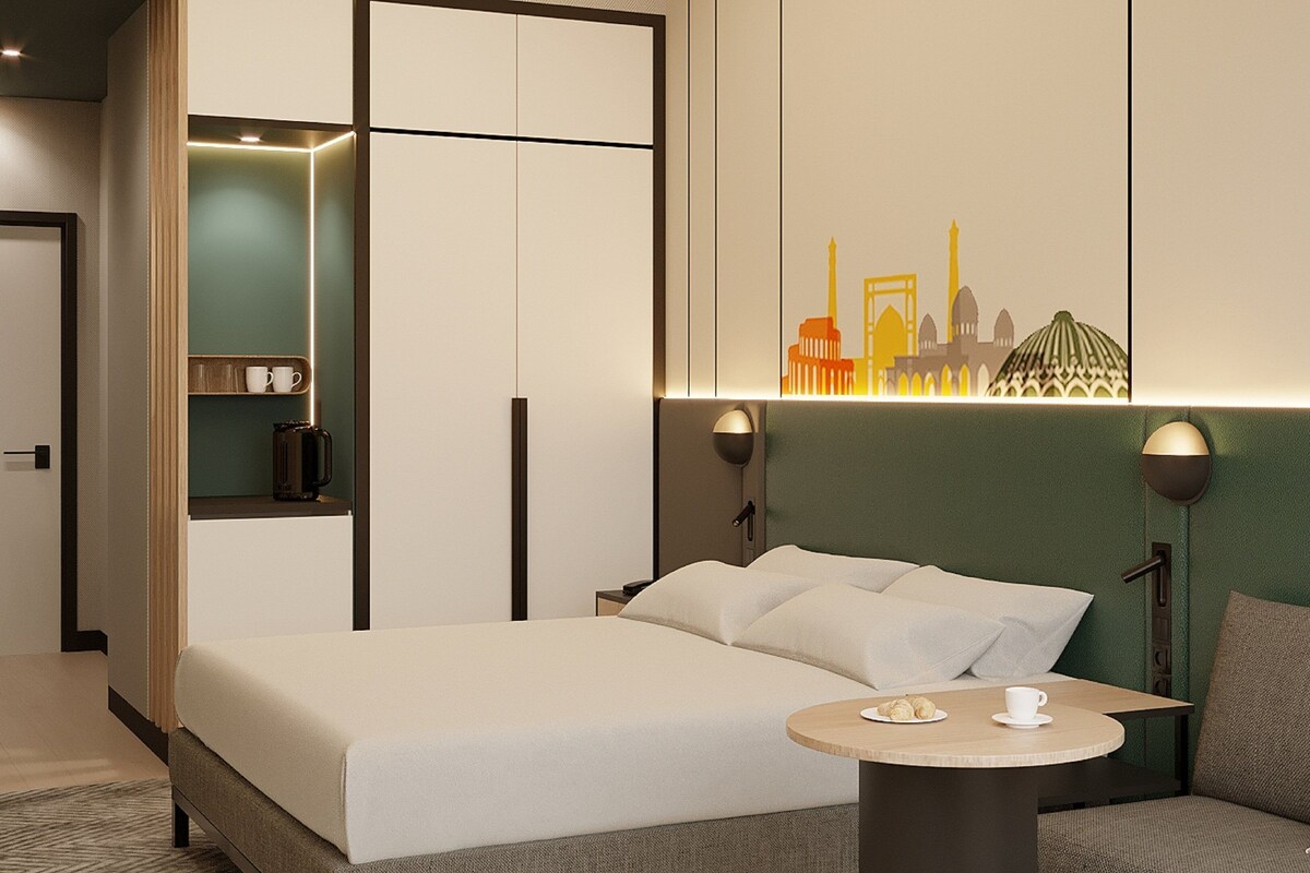 A modern hotel room with a bed, a small round table, a coffee machine, and a wall-mounted artwork.